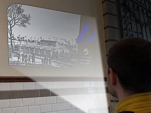 multimediale Leipzig Stadtführung: der Guide, dessen Kopf man rechts unten in der Ecke von hinten sieht, projiziert mit einem mobilen Beamer ein Bild an die Wand, welches eine historische Eisenbahn zeigt
