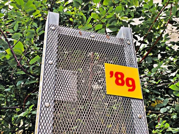 Wendezeit 1989 - Friedliche Revolution - zu sehen eine Infostele aus Metall mit einem engmaschigen Gitter vorn, rechts oben daran eine rote Zahl 89 auf gelbem Grund. Im Hintergrund ein grüner Busch oder Baum.