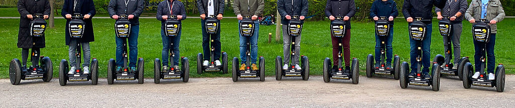 Gruppenfoto mit 12 Personen auf Segways nebeneinander am Rand eines Weges vor einer Wiese. Die Personen sind ab den Oberkörpern abwärts erkennbar (ohne Köpfe), da der Fokus auf den Segways liegt, mit denen Touren in Leipzig durchgeführt werden.